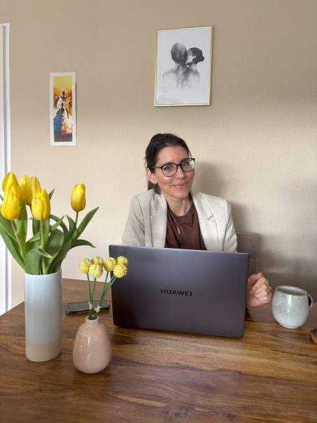 Eine Person mit Brille sitzt an einem Tisch, umgeben von Blumen und Laptop.