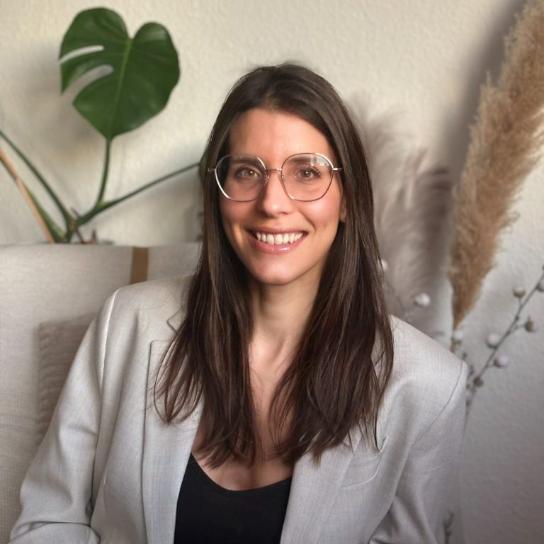 Bild bis Brust, sitzend auf Stuhl, lächelnd offener Mund, offene Haare, goldene Brille, beiger Blazer, im Hintergrund Monstera Pflanze 
