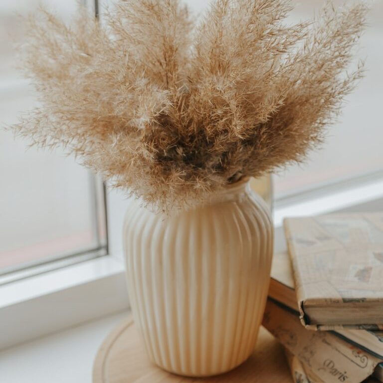 Beige Trockenblumen in einer gerippten Vase auf einem Fensterbrett.